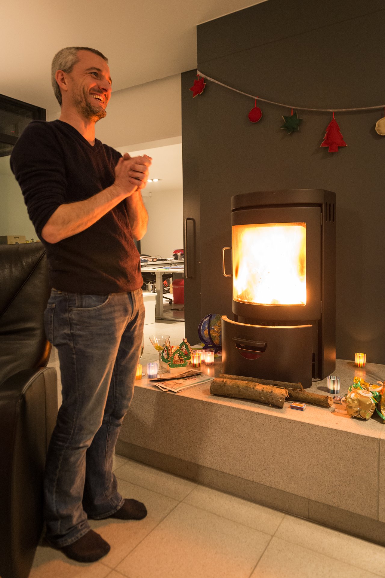 A man smiles and warms his hands near a fireplace, surrounded by candles and festive decorations.