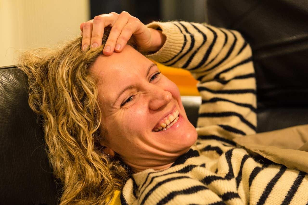 A woman with curly hair and a striped sweater laughs while reclining on a couch at a New Year's Eve party.