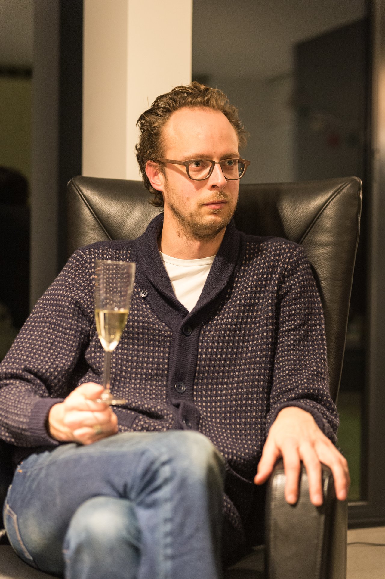A man sits in a chair, holding a glass of champagne and looking to the side.
