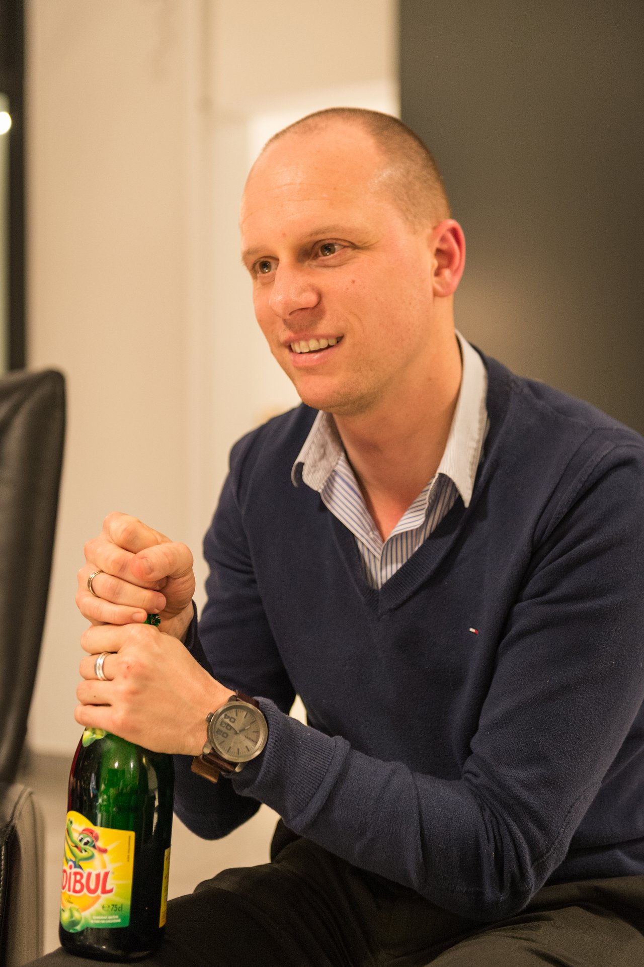 A man in a blue sweater is smiling while opening a bottle at a New Year's Eve party.