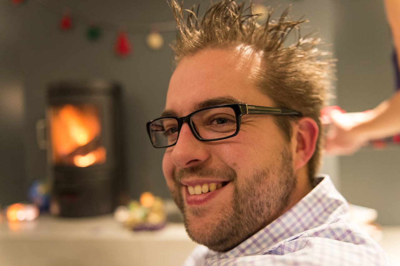 A smiling man with glasses and spiky hair sits indoors while someone styles his hair in the background.