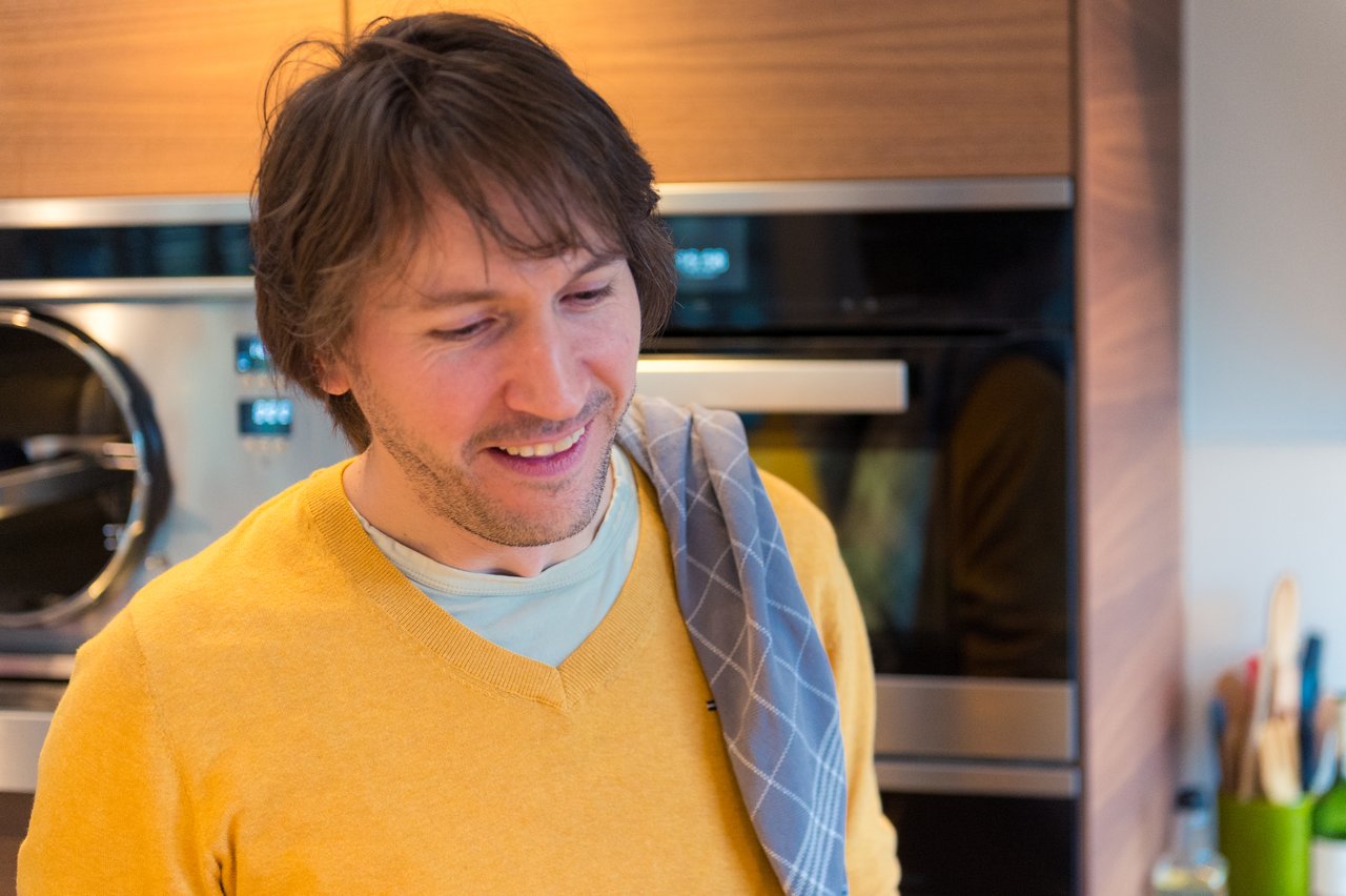 A man in a yellow sweater smiles in a kitchen with a towel over his shoulder.