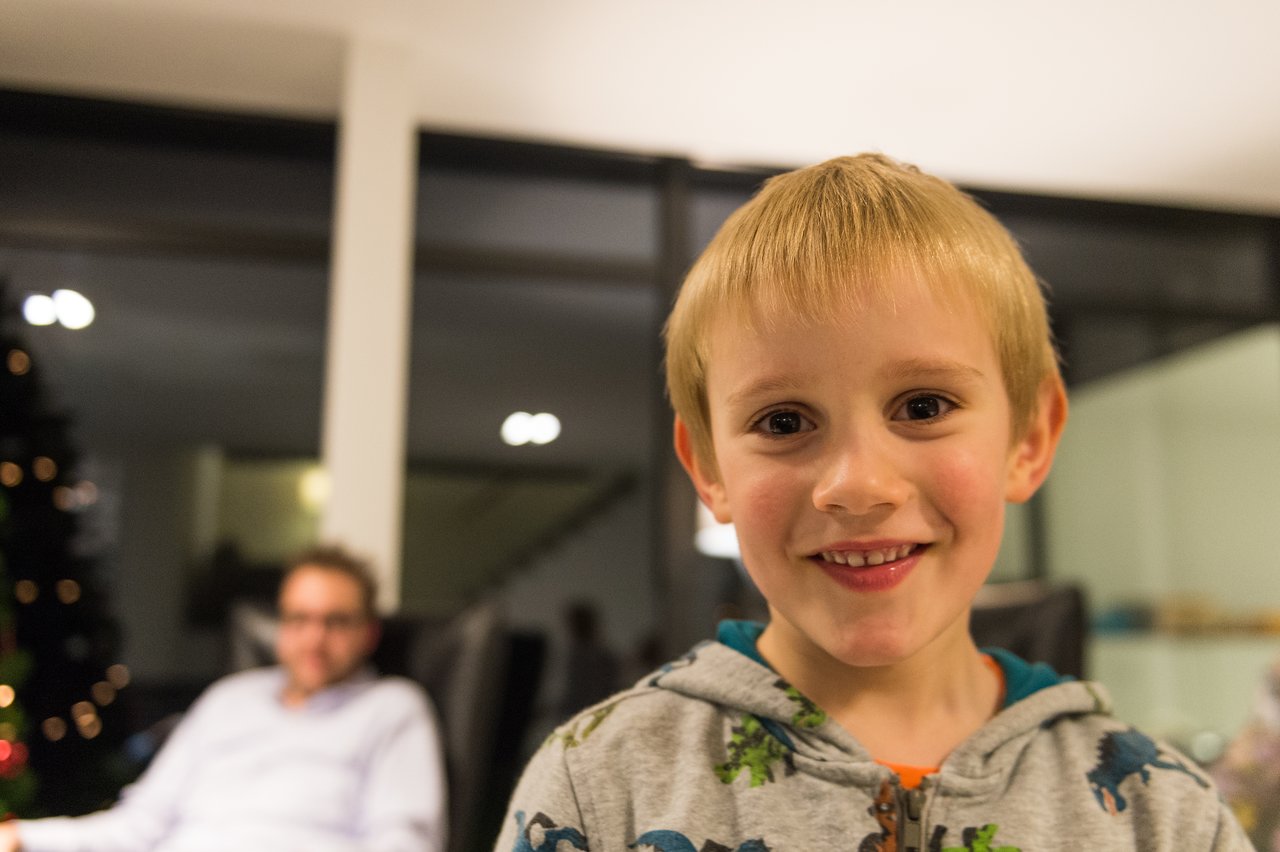 A smiling child in a dinosaur hoodie stands in the foreground, with an adult sitting in the background.