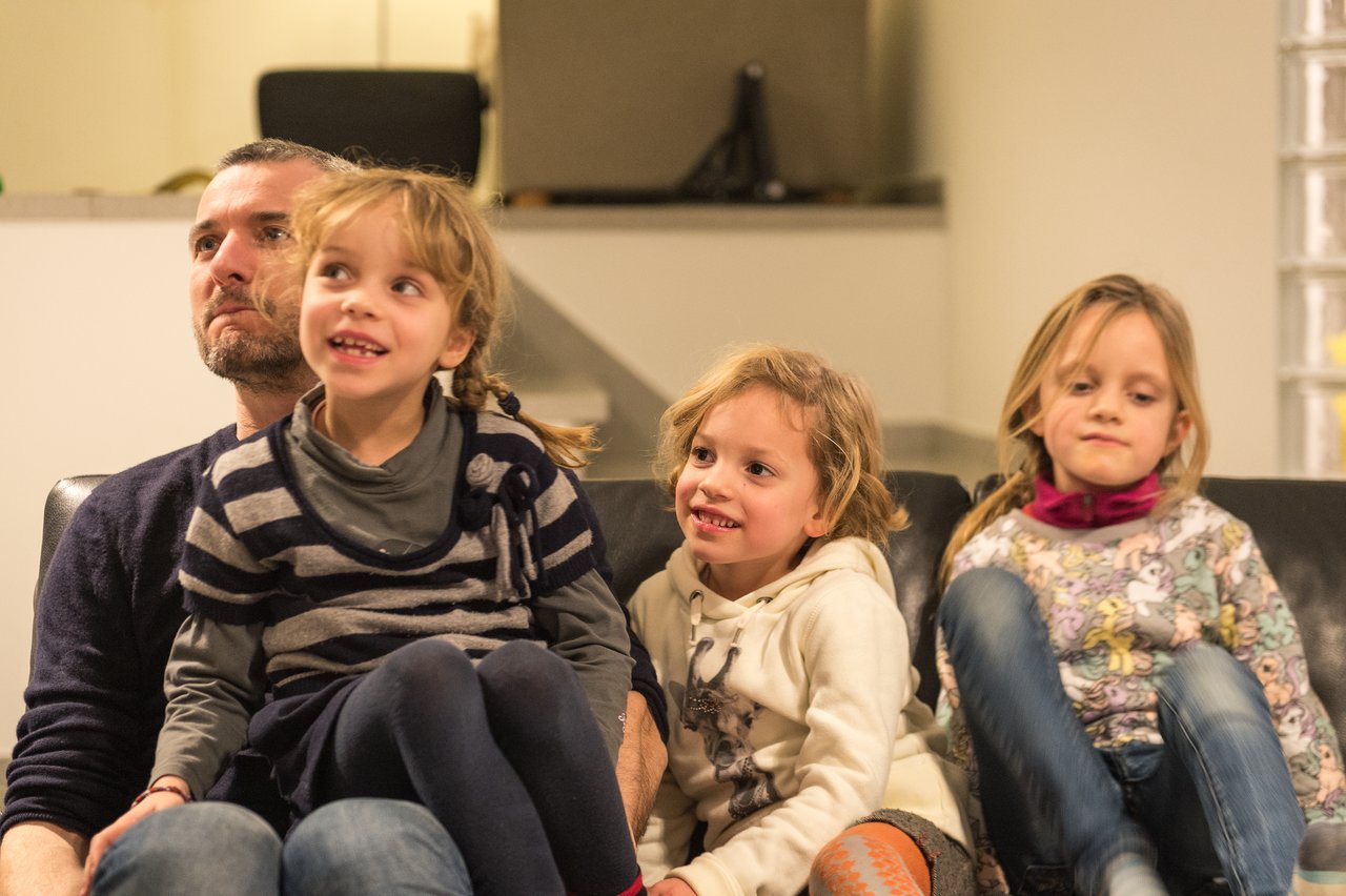 A man sits on a couch with three young children, two on his lap, all appearing engaged and happy.