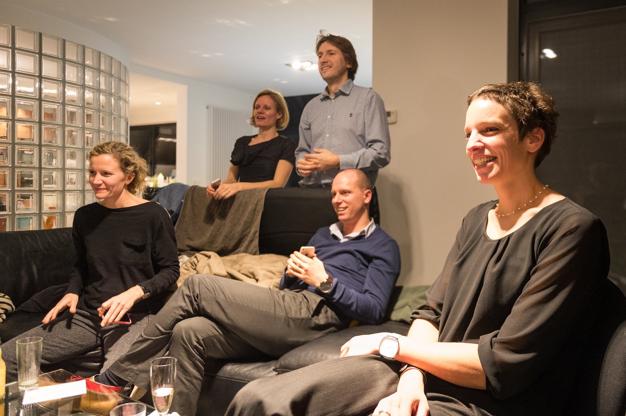 A group of people sitting and standing in a living room, smiling and engaged in conversation at a New Year's Eve party.