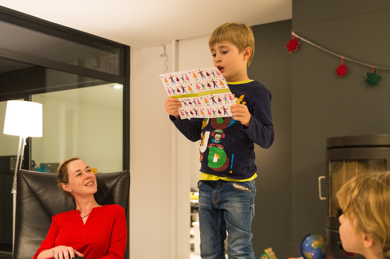 A young boy stands on a chair, reading from a colorful paper, while others watch and smile.