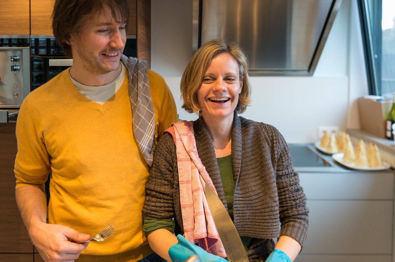 A smiling woman in a kitchen holds a large knife, while a man beside her laughs and holds a fork.