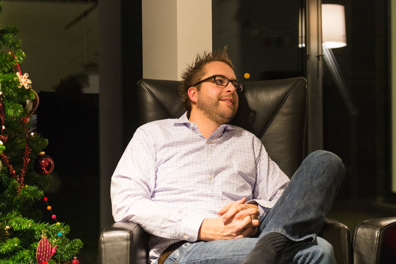 A man in glasses and a button-up shirt sits in a chair, smiling and relaxing at a New Year's Eve party.