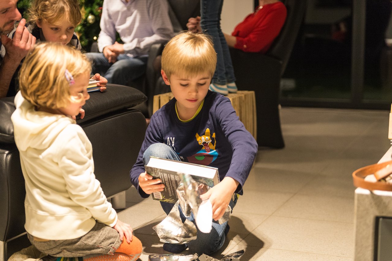 A young boy sits on the floor, unwrapping a gift, while a girl watches closely beside him.