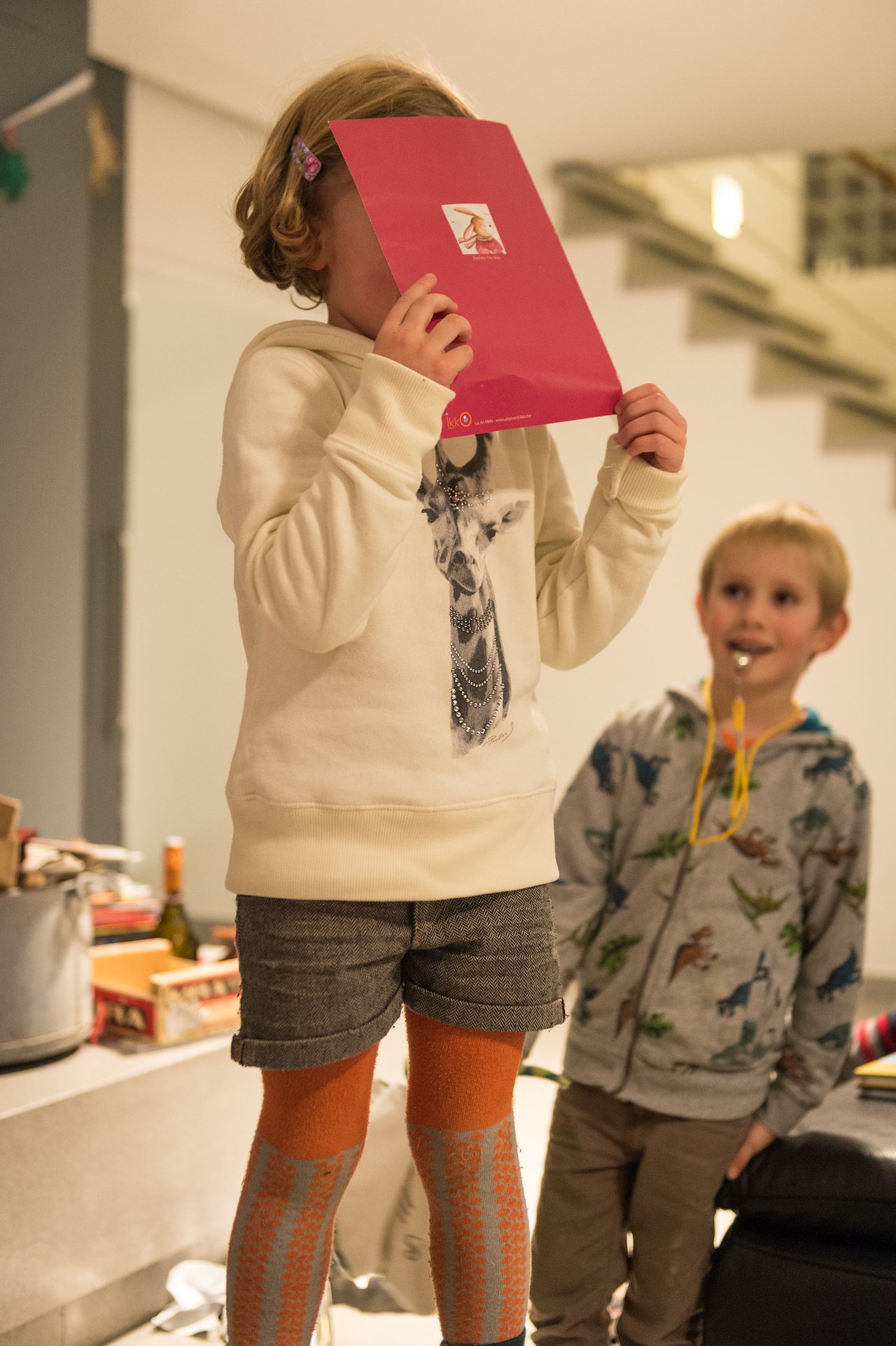 A child holds a red card in front of their face while another child watches with a party blower.