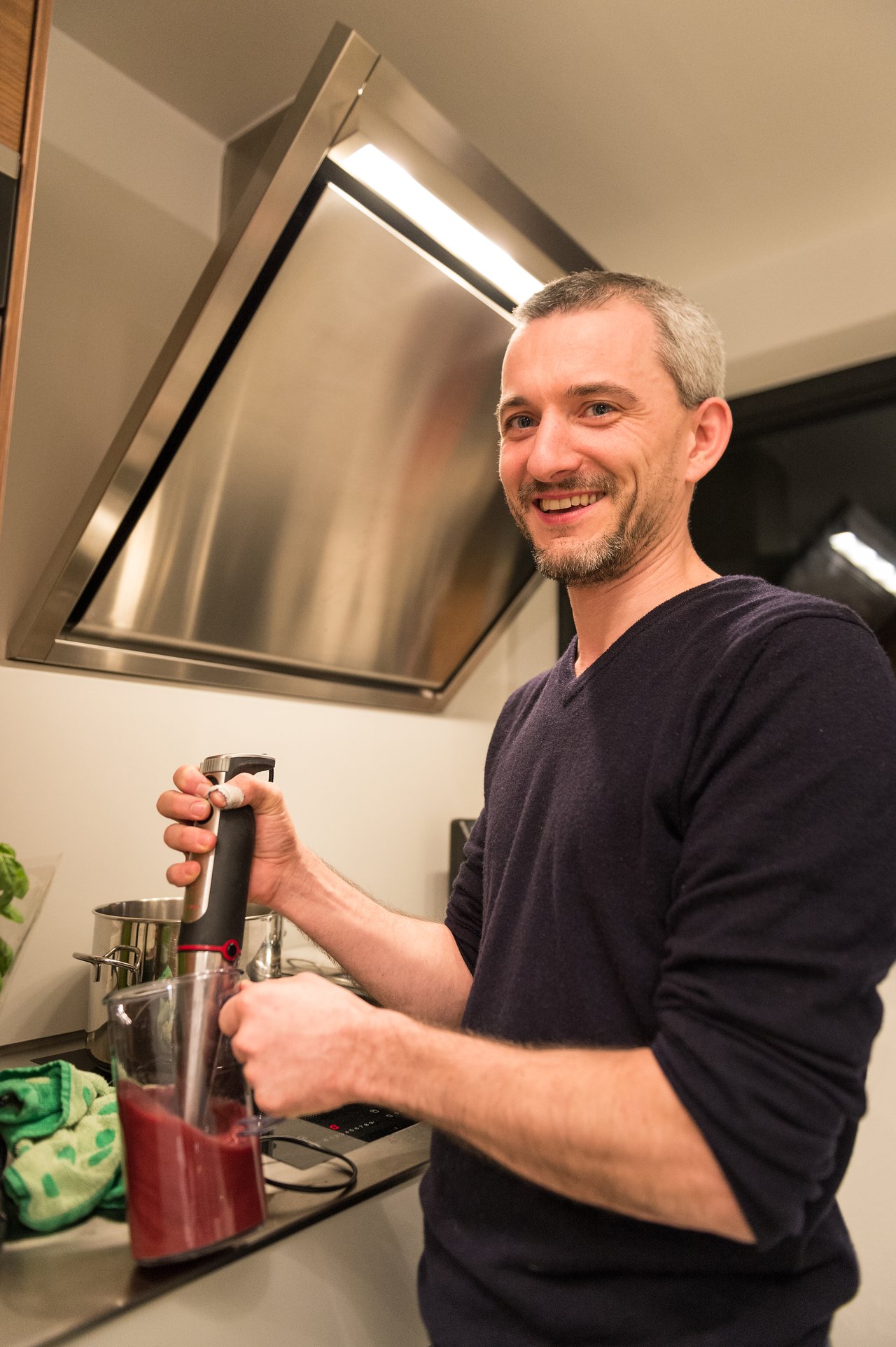 A man smiles while using a hand blender to mix a red drink in a kitchen.