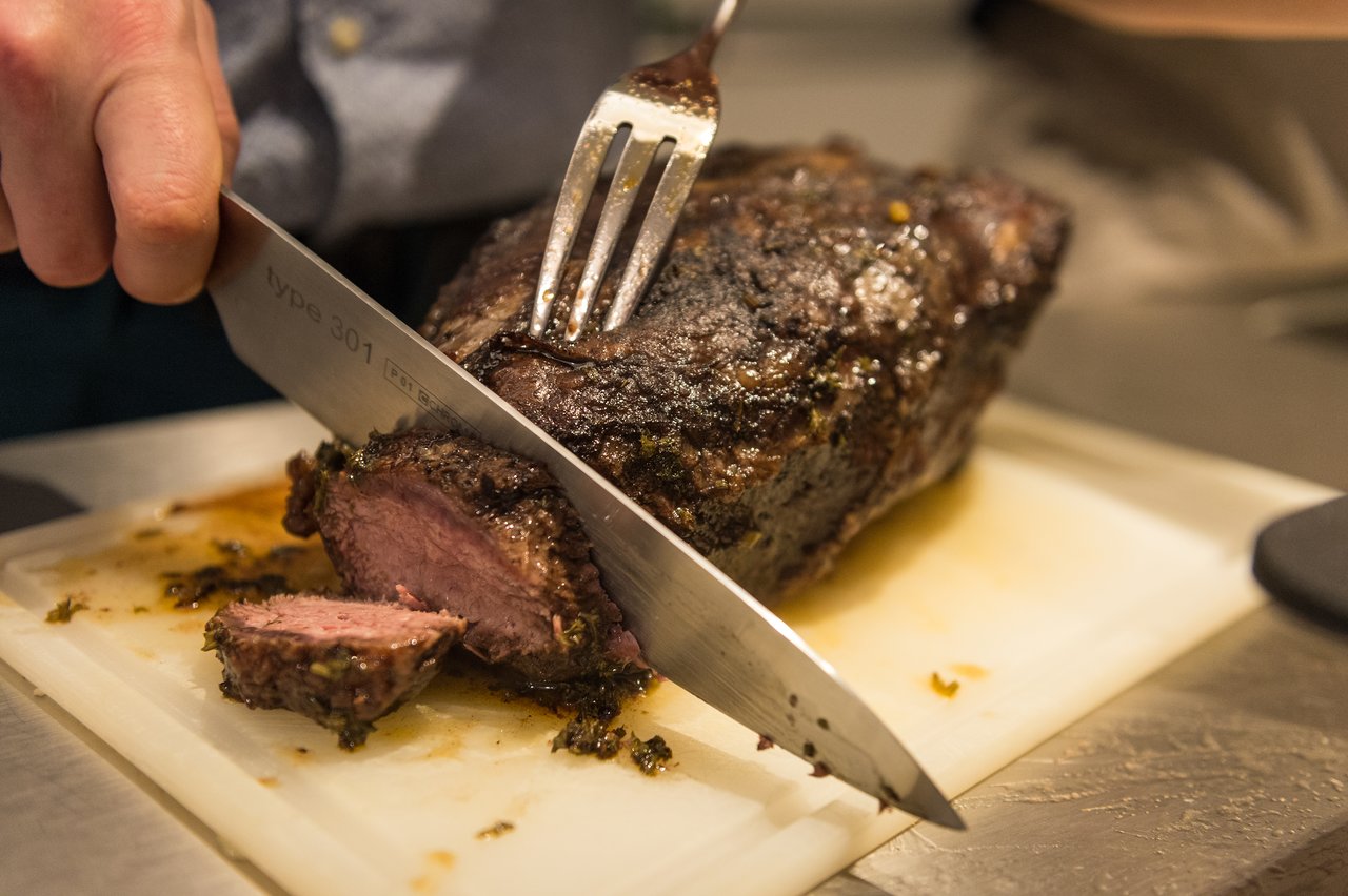 A person slices a roasted piece of meat on a cutting board using a knife and fork.
