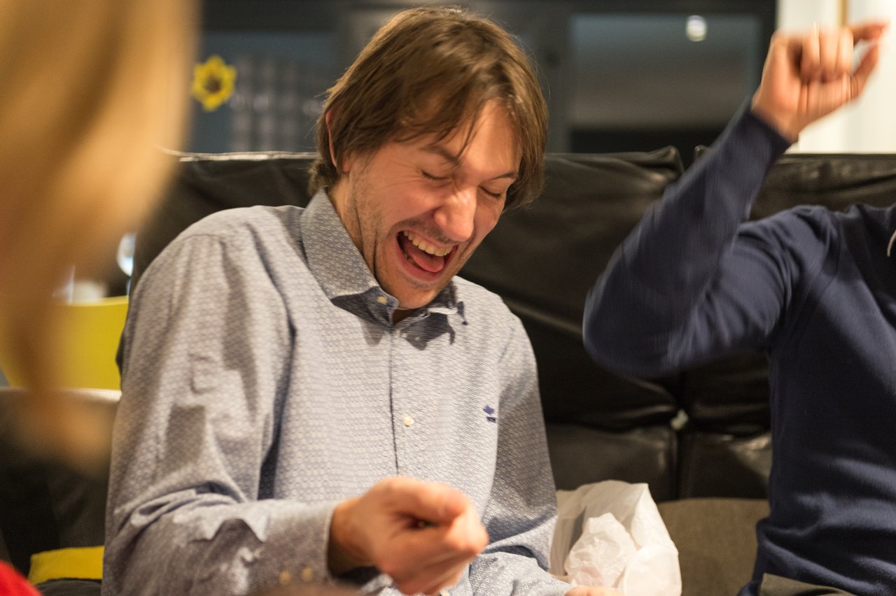 A man in a light-colored shirt laughs joyfully while sitting on a couch at a New Year's Eve party.