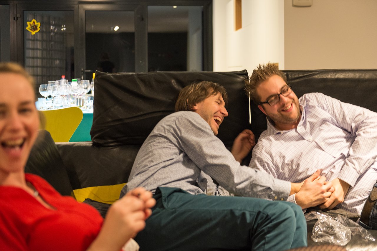 Three friends laughing on a couch, with two men playfully leaning on each other and a woman smiling in front.