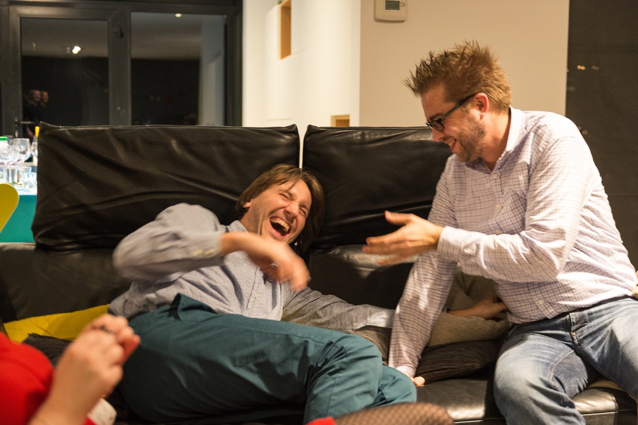 Two men laughing on a couch, one leaning back while the other gestures playfully.