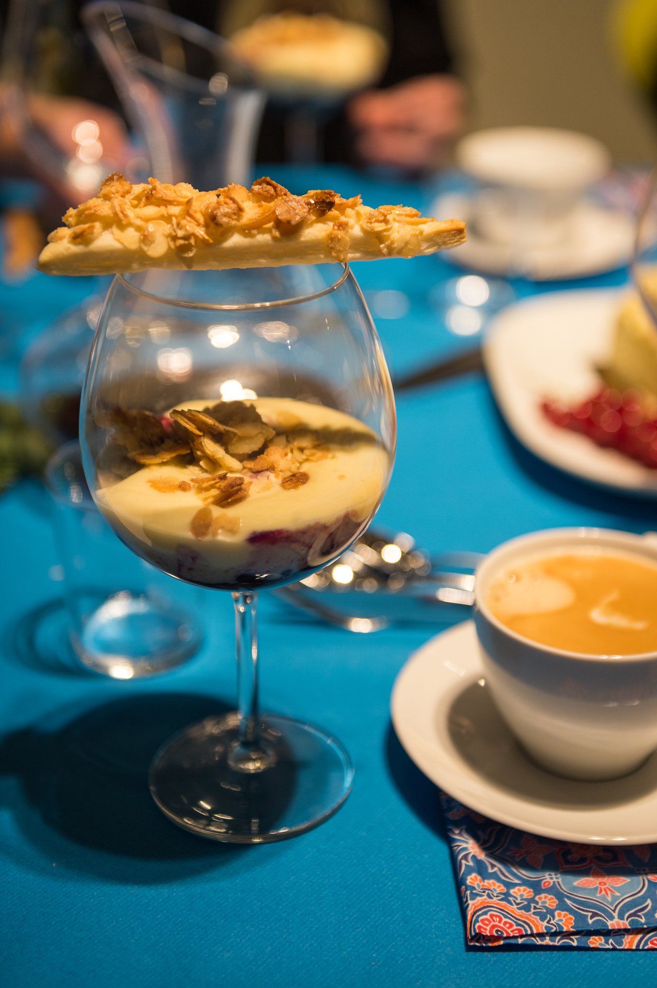 A dessert glass with layered cream and fruit, topped with a pastry, next to a cup of coffee.