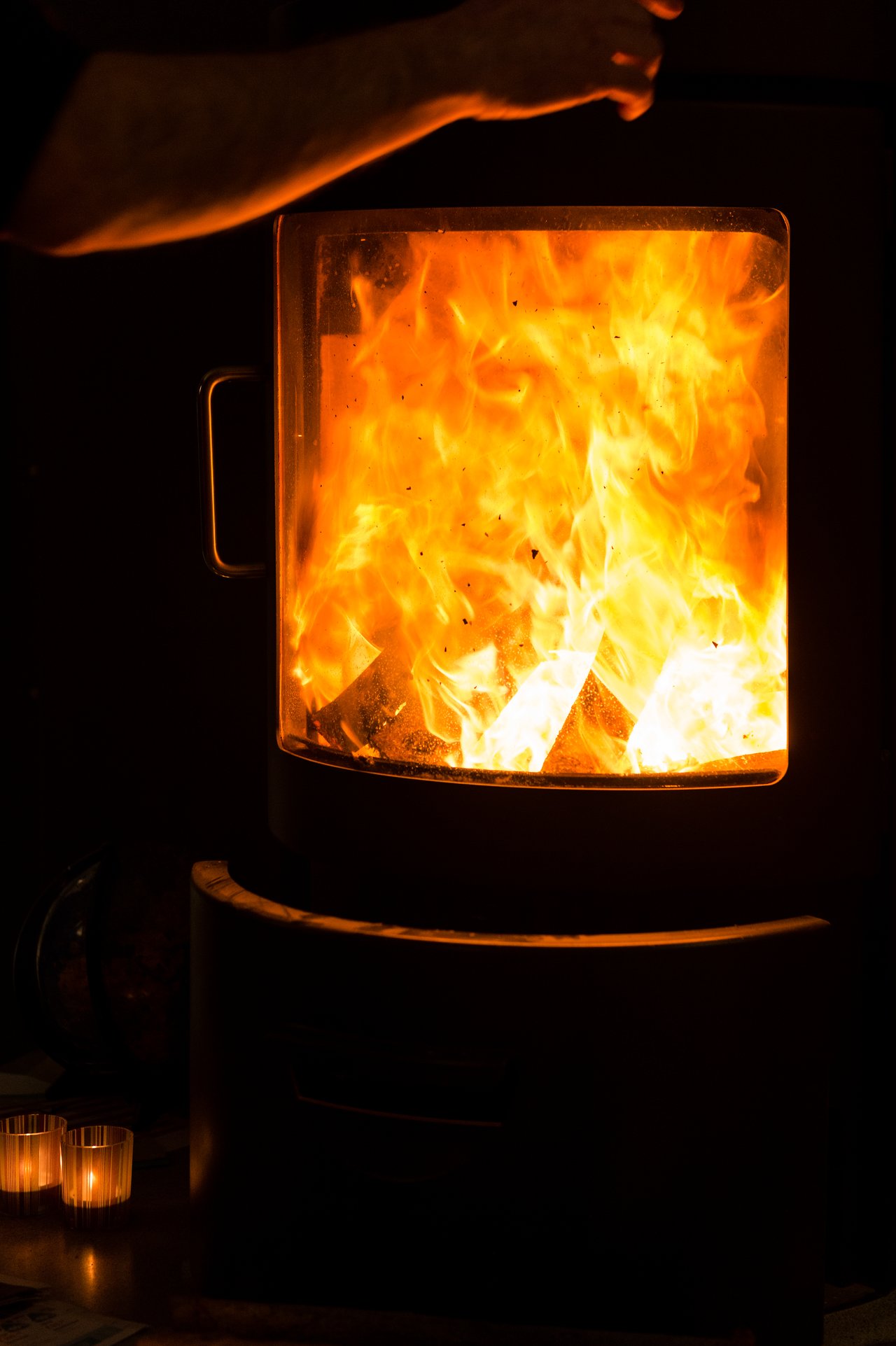A person adjusts a wood-burning stove with bright flames inside, while small candles glow nearby.