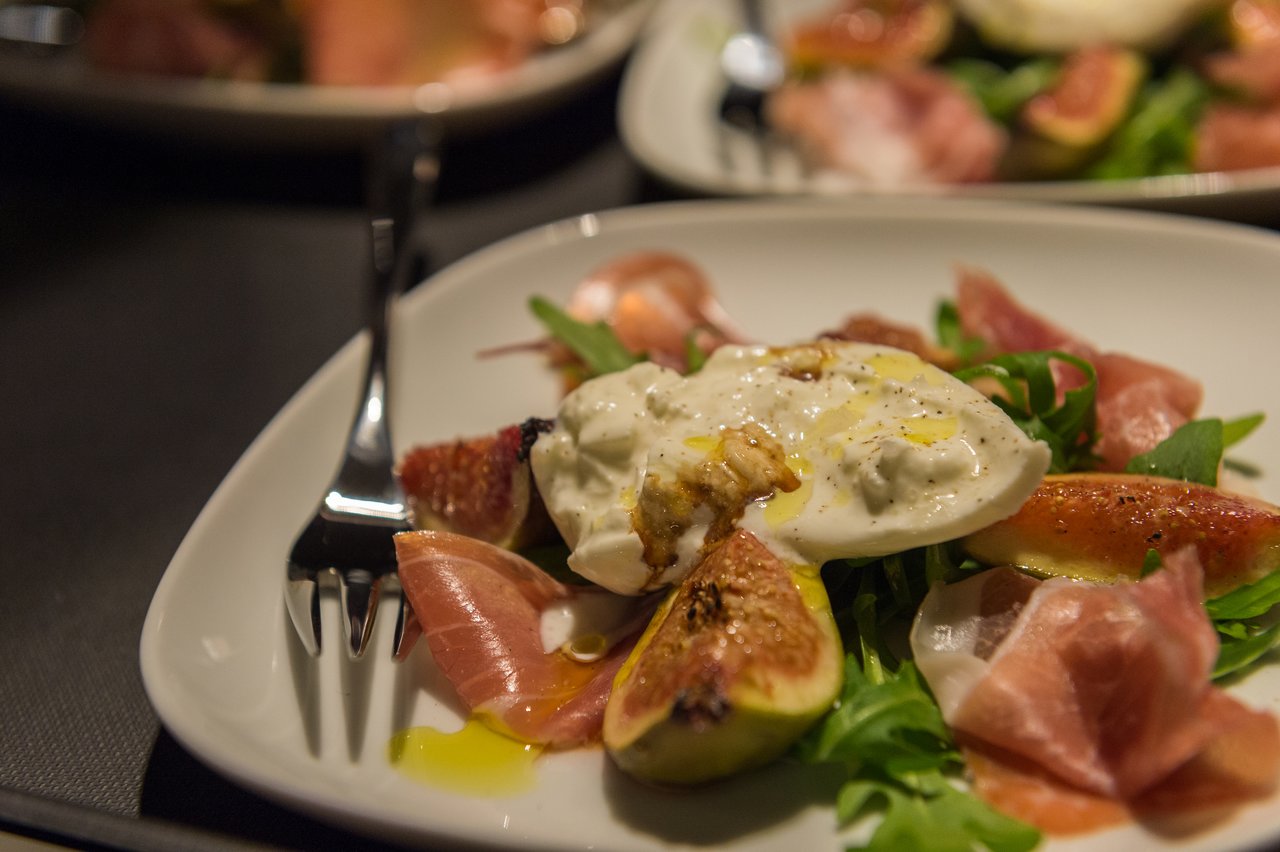 A plate of salad with prosciutto, fresh figs, arugula, and creamy burrata cheese, drizzled with olive oil.