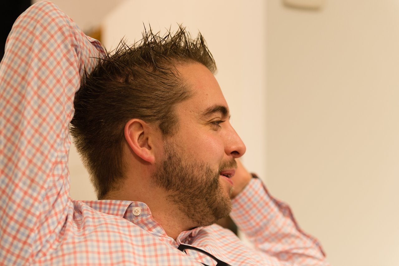 A man in a checkered shirt leans back with his hands behind his head, appearing relaxed at a party.