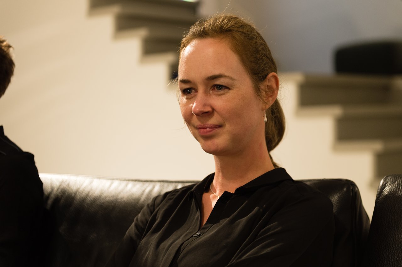 A woman in a black shirt sits on a black couch, smiling slightly while looking at something off-camera.