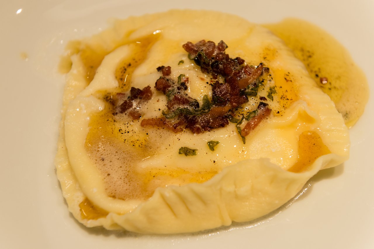A plated raviolo topped with crispy bacon, herbs, and melted butter sauce.