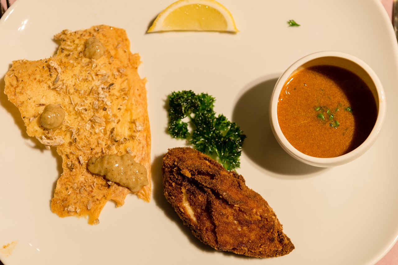 A plated meal with fried fish, a crispy flatbread, a small bowl of sauce, parsley, and a lemon wedge.