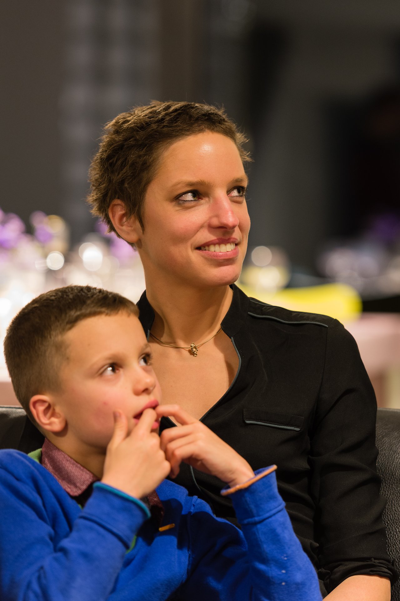 A woman in a black shirt smiles while sitting with a young boy in a blue sweater.