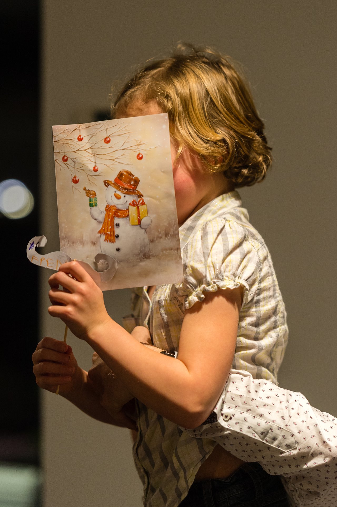 A child holds up a festive snowman card, partially covering their face, during a New Year's Eve party.