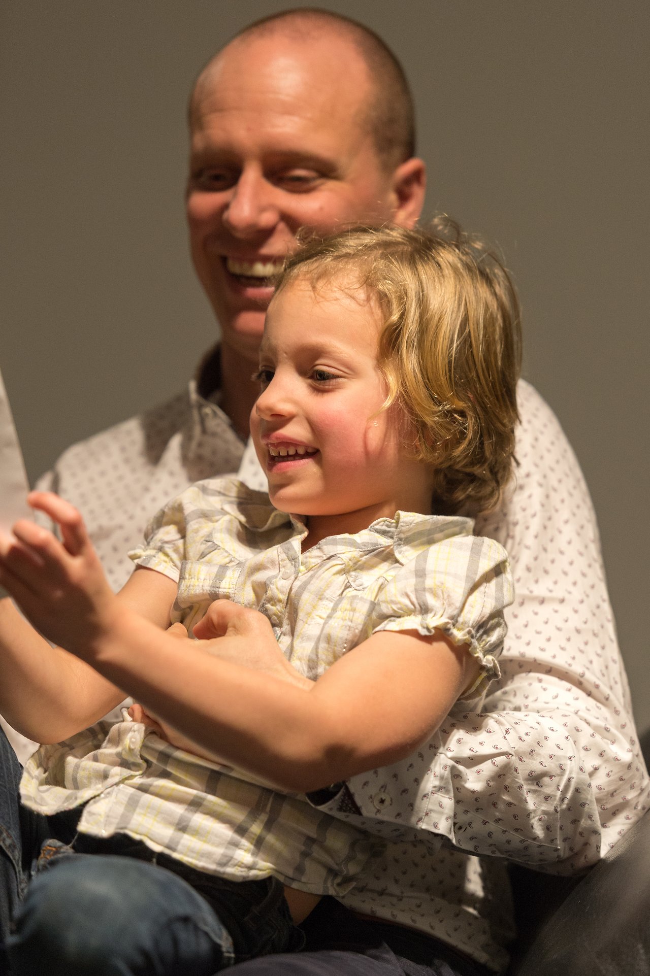 A smiling child sits on an adult's lap, holding something and looking at it with excitement.