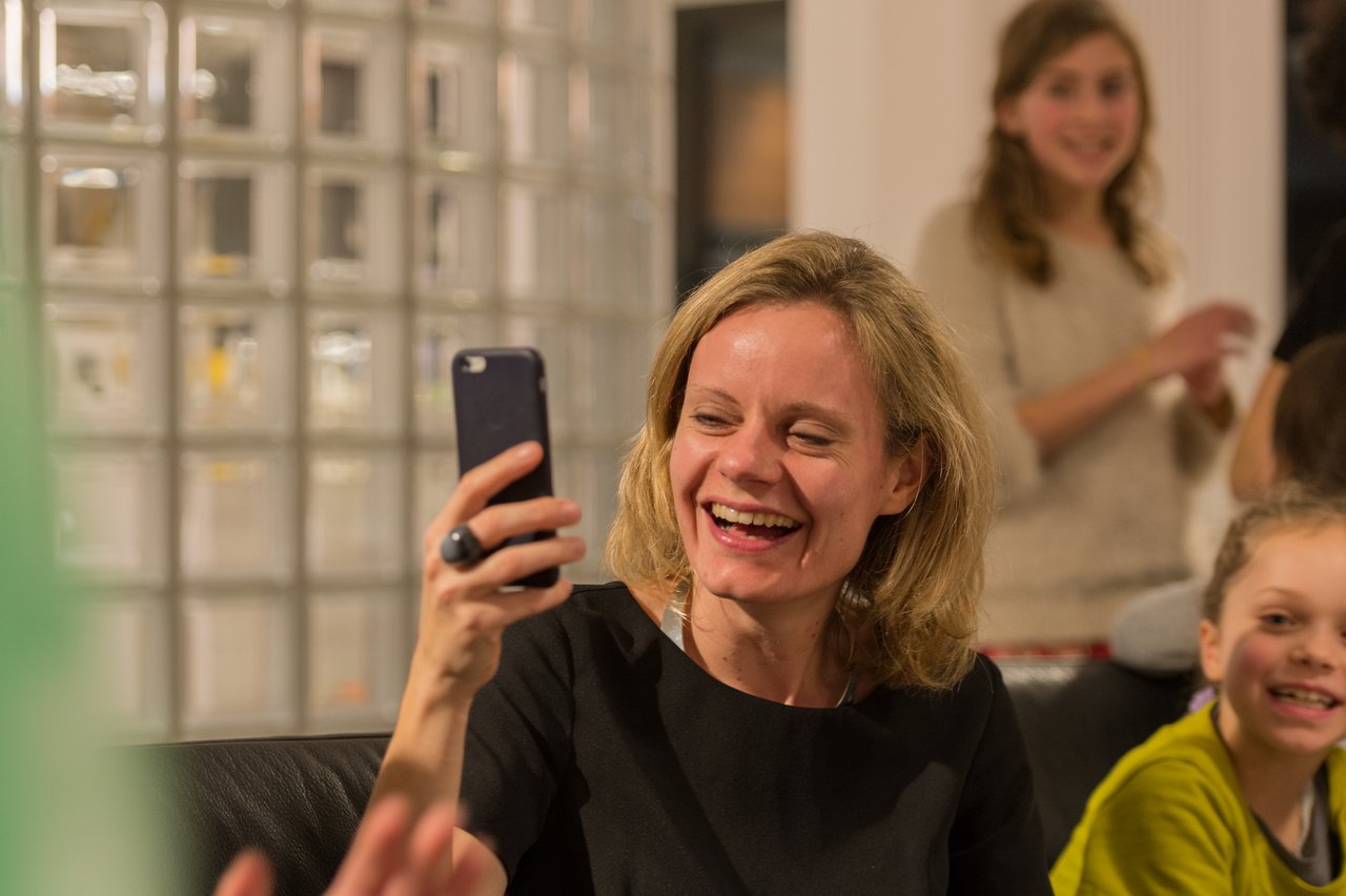 A smiling woman in a black dress holds up her phone, surrounded by people at a lively New Year's Eve party.