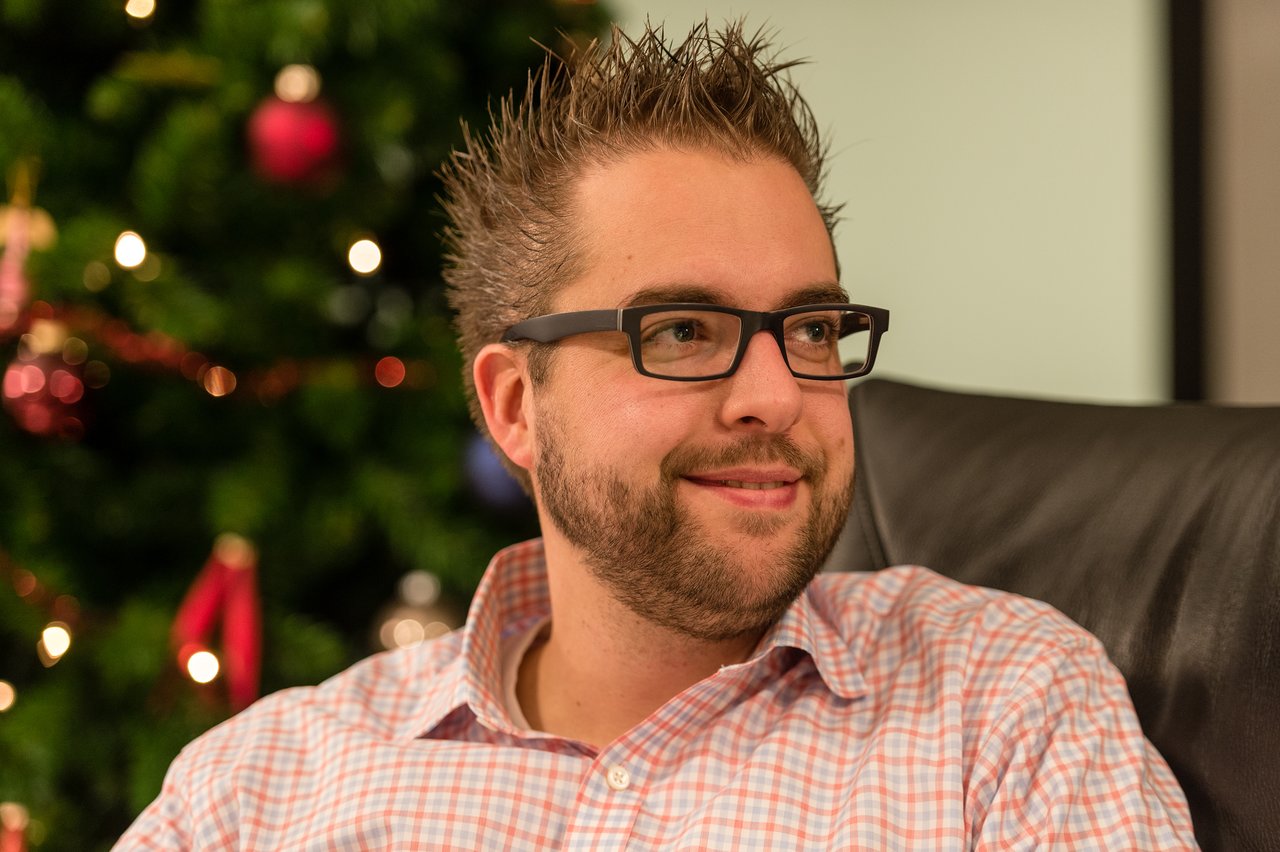 A man wearing glasses and a checkered shirt smiles while sitting in a chair, with a decorated tree behind him.