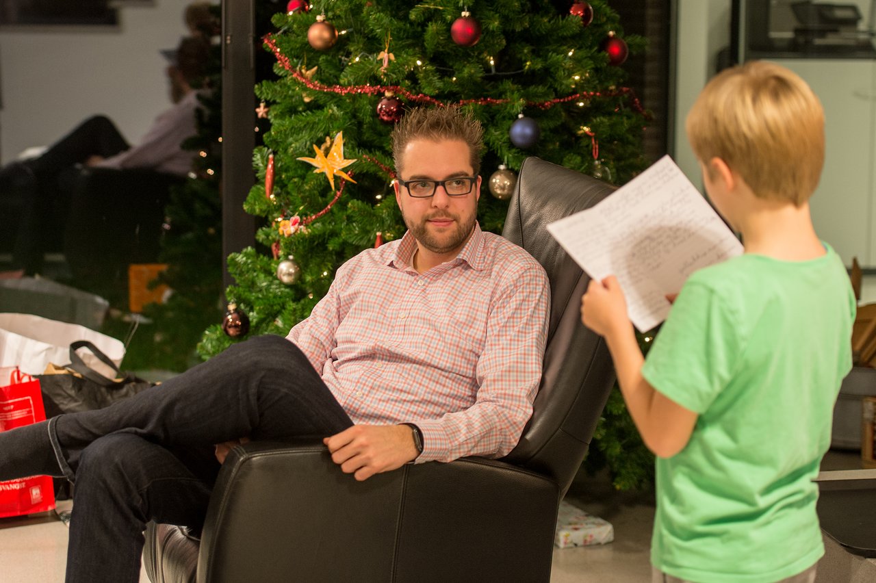 A child in a green shirt reads from a paper while an adult listens, sitting near a decorated Christmas tree.
