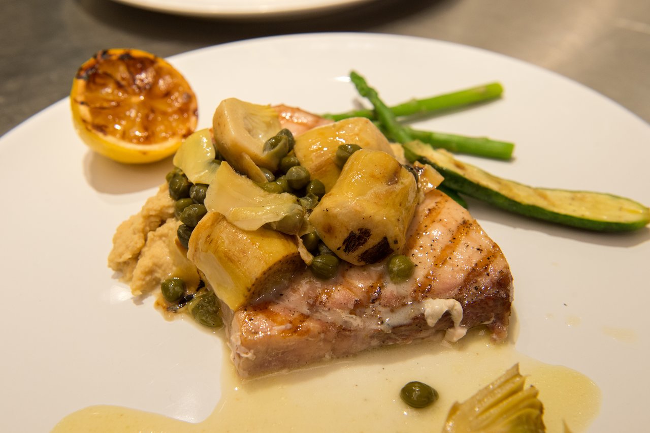 A plated dish with grilled fish, artichokes, capers, sauce, and vegetables, served with a charred lemon on the side.