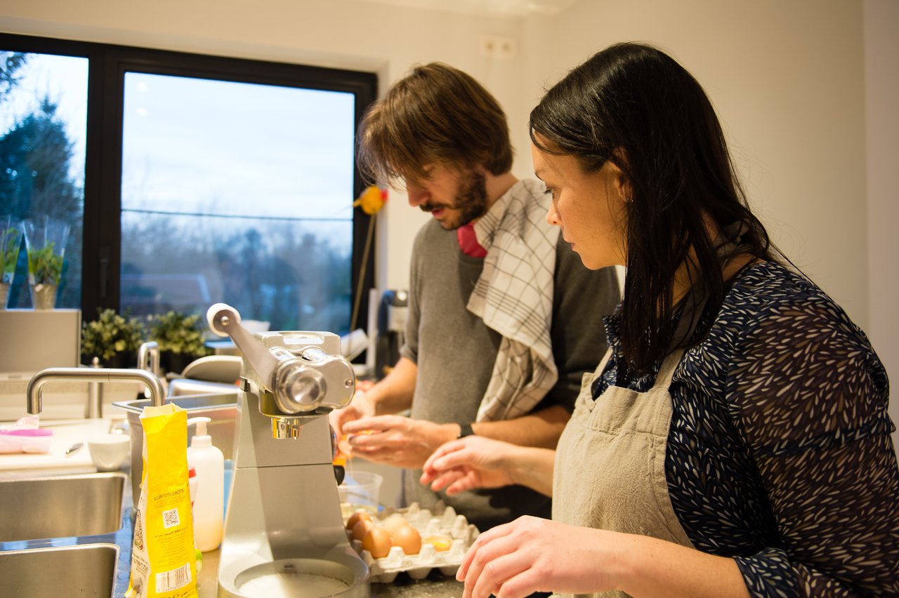 Two people in a kitchen preparing food, cracking eggs and using a pasta machine.