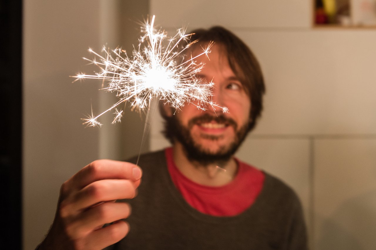 A man holds a lit sparkler and smiles, celebrating at a New Year's Eve party.