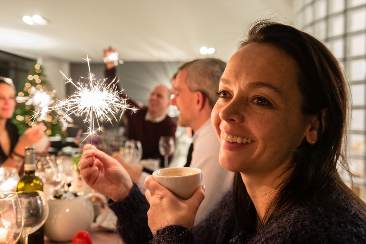 A smiling woman holds a cup while people around her celebrate with sparklers at a New Year's Eve party.
