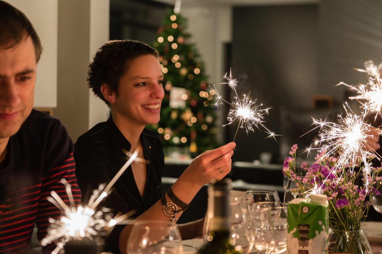 People celebrating New Year's Eve, holding sparklers at a table with drinks and decorations.