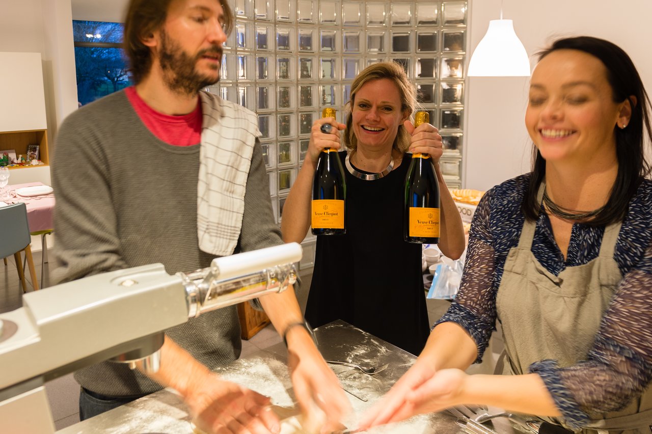 A woman holds two bottles of champagne while two people prepare food in a kitchen.