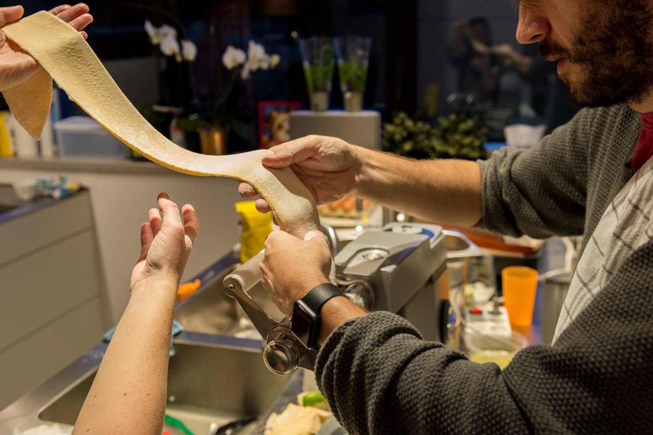 Two people making fresh pasta, feeding dough through a pasta machine in a kitchen.