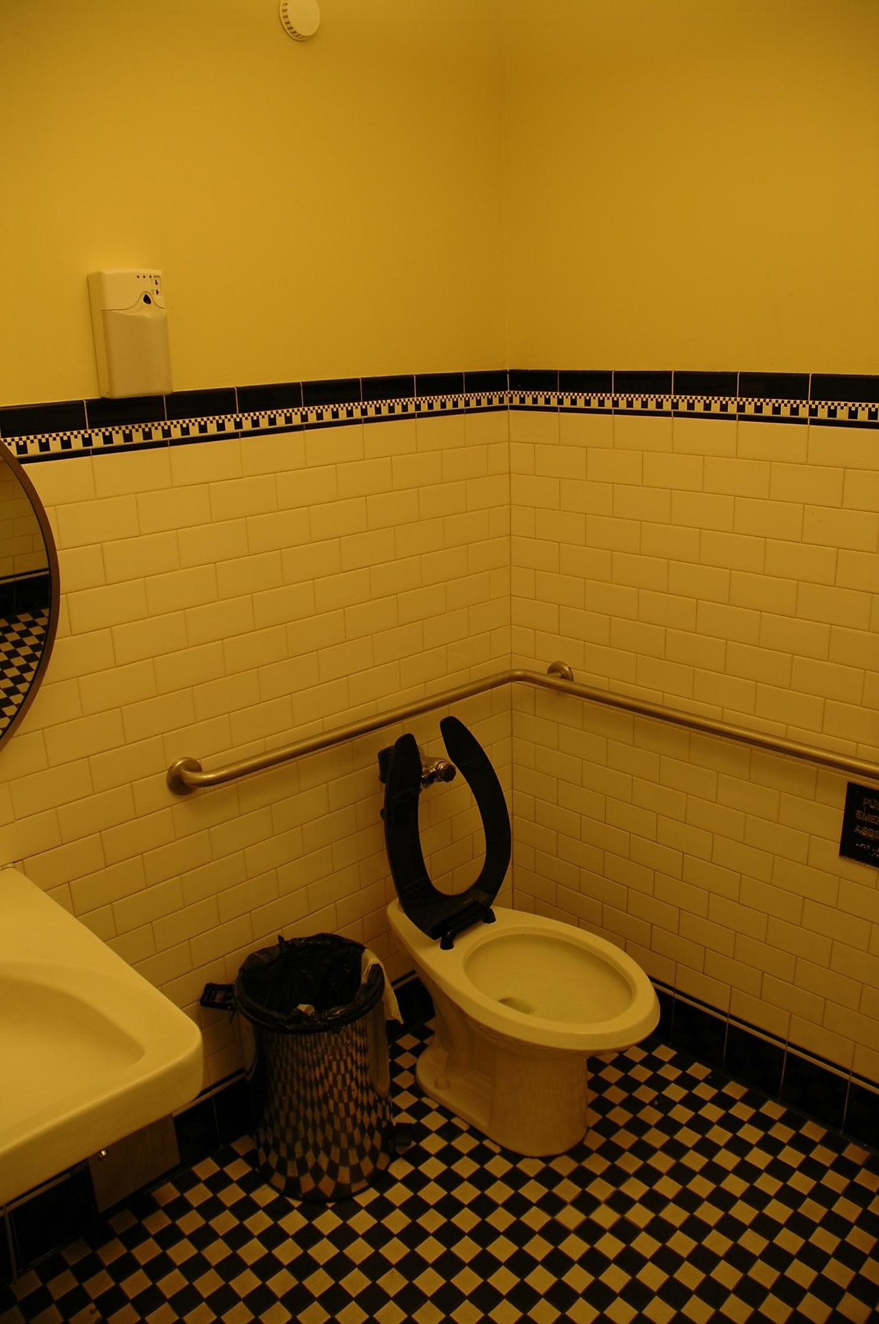 A public restroom with a toilet, open black seat, metal trash bin, and grab bars against tiled walls.