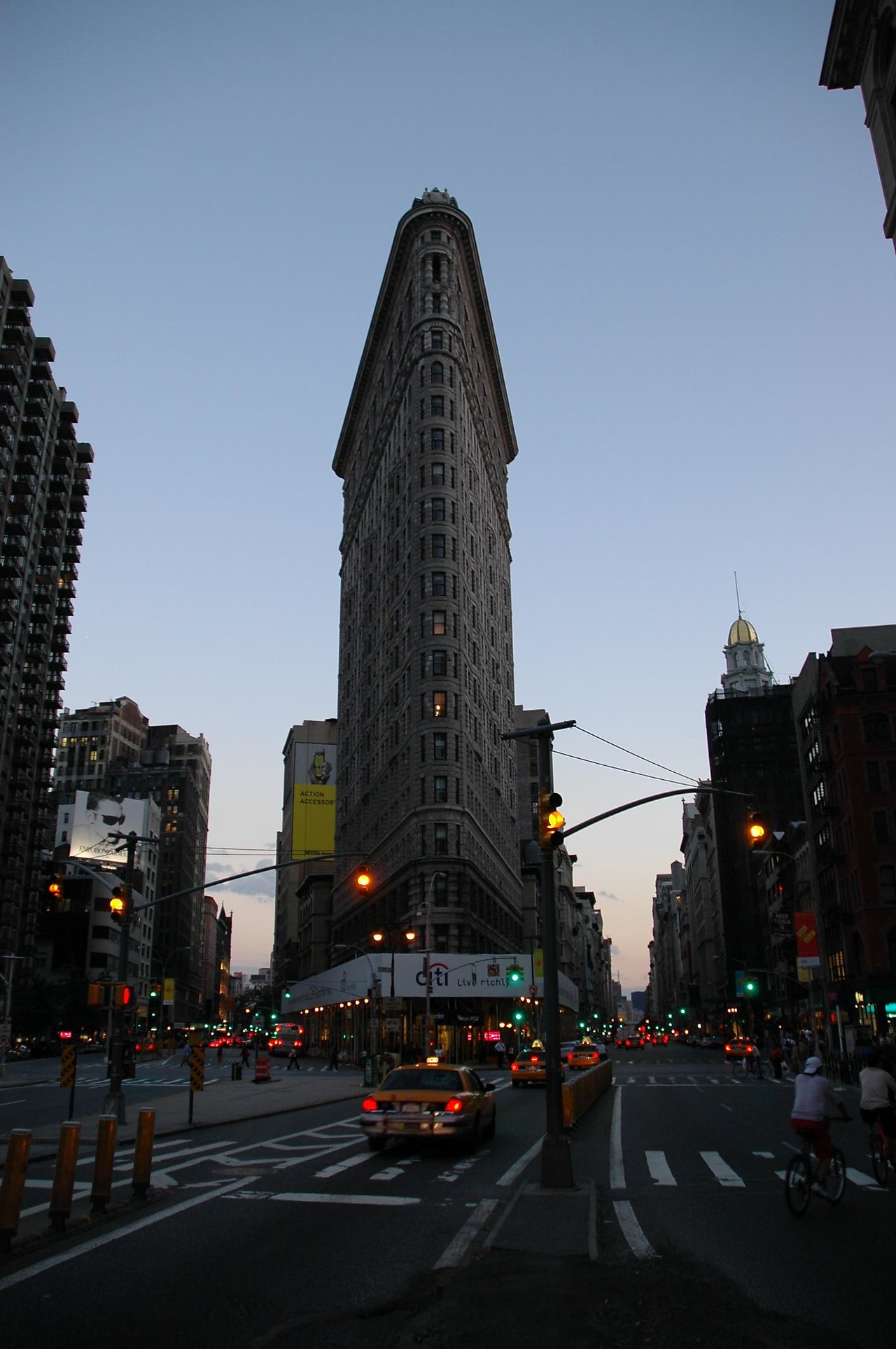 The Flatiron Building stands tall at dusk, with taxis and cyclists moving through the busy New York City streets.