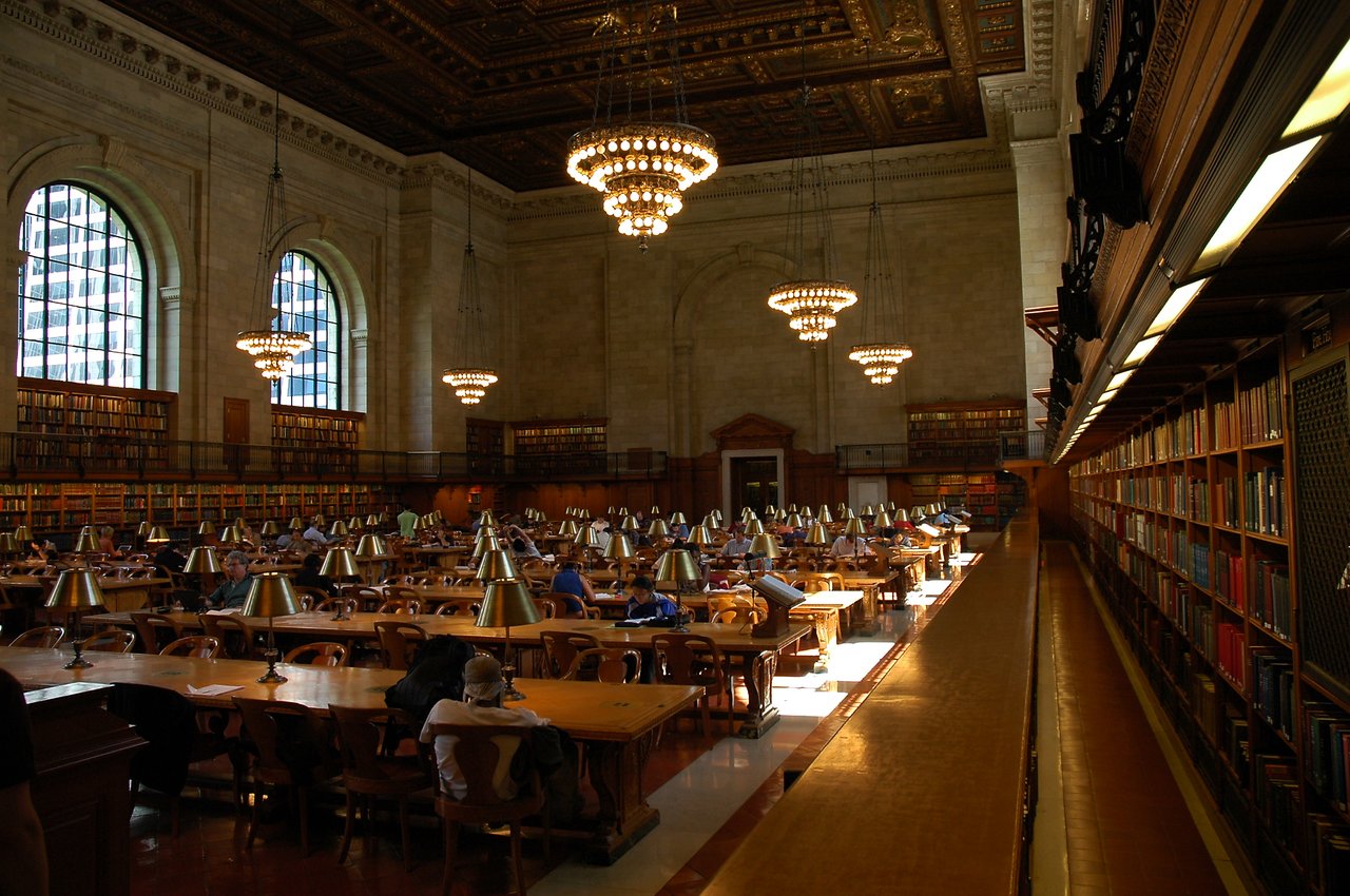 A large reading room in a public library with people studying at wooden tables under hanging chandeliers.