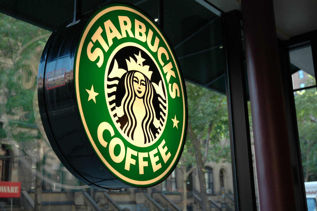 A bright Starbucks Coffee sign hangs outside a café, with a city street visible through the window.
