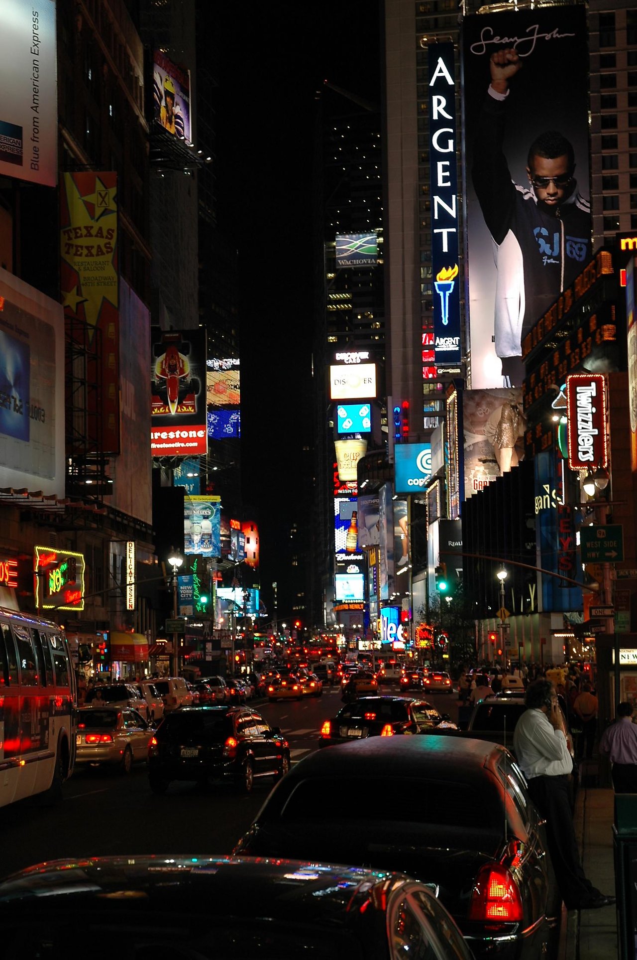 Busy Times Square at night with bright billboards, heavy traffic, and people standing near parked cars on the sidewalk.