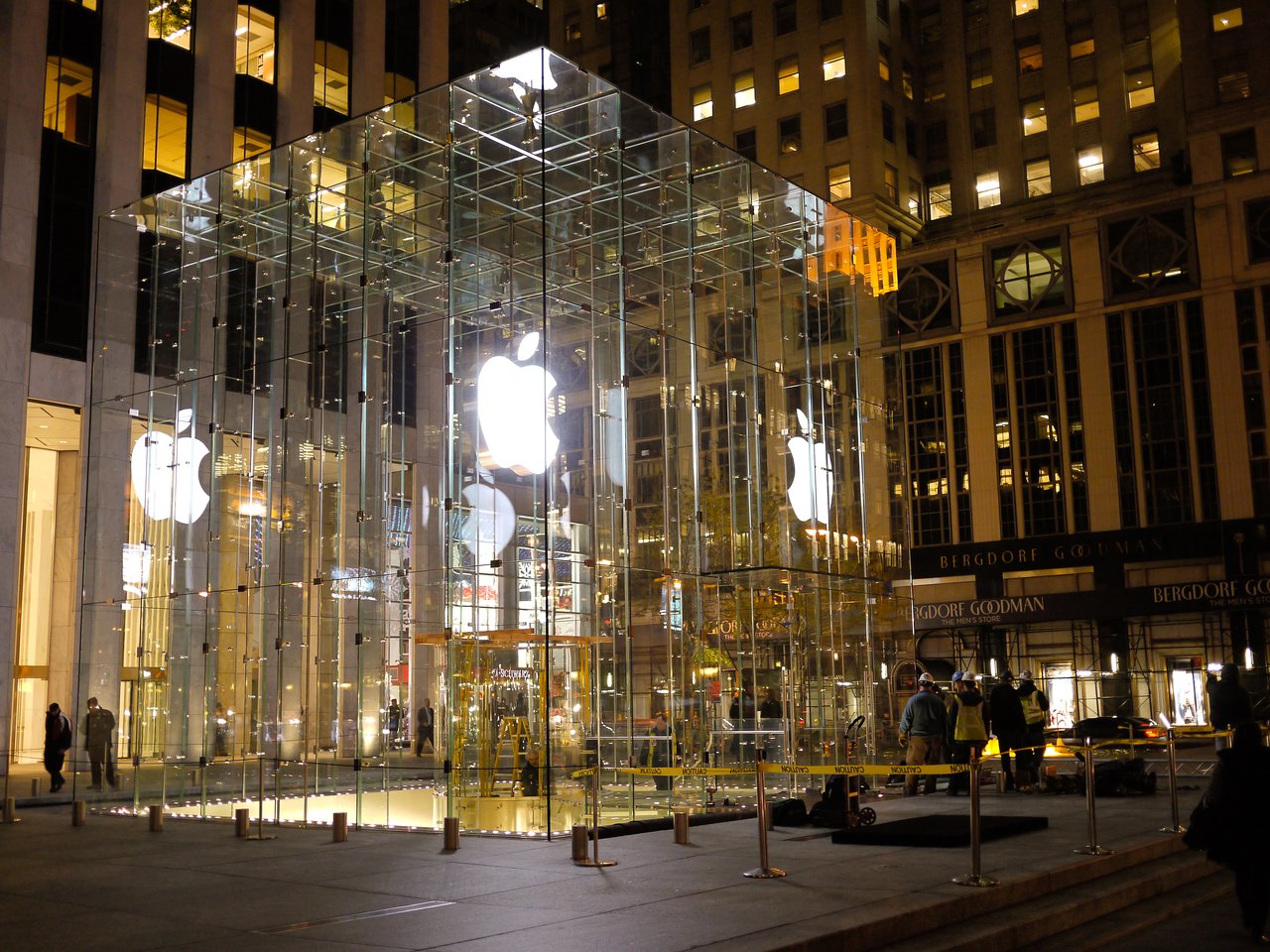 Glass cube entrance to an Apple Store at night, with illuminated Apple logos and people walking nearby.
