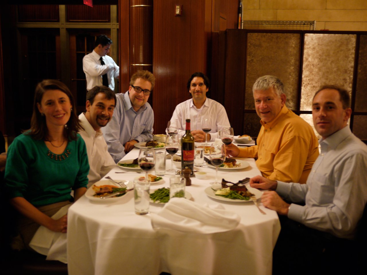 A group of six people sitting at a restaurant table, enjoying a meal and conversation during a Drupal Association meeting.