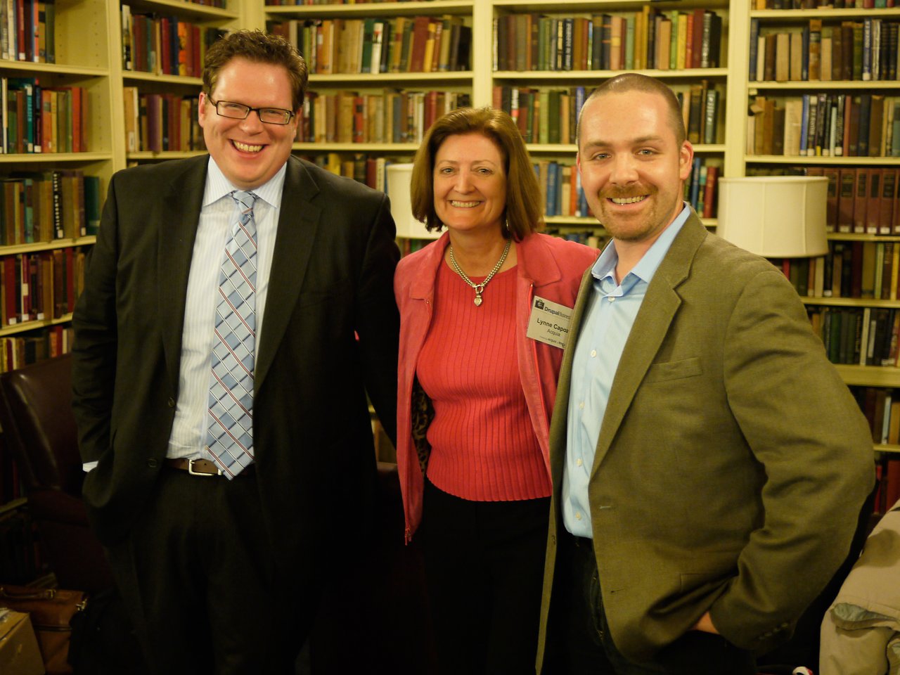 Three people smiling and posing at the Drupal Business Summit reception, standing in front of bookshelves.