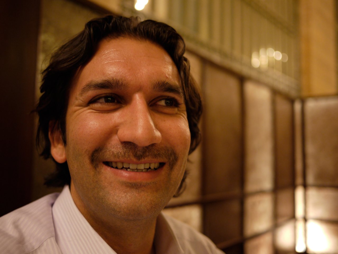 A man with dark hair and a mustache smiles while looking slightly to the side in a warmly lit setting.