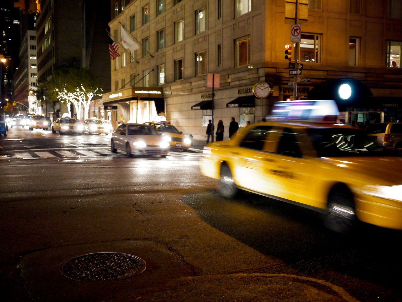 A yellow taxi moves through a busy city street at night, with other cars and pedestrians in the background.