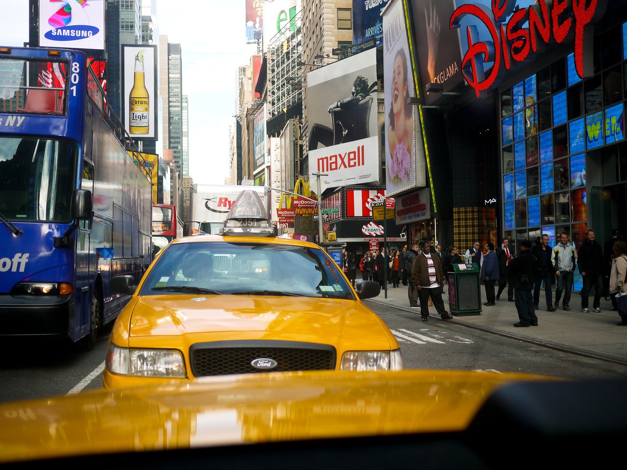 A yellow taxi in traffic at Times Square, surrounded by buses, pedestrians, and bright advertising billboards.